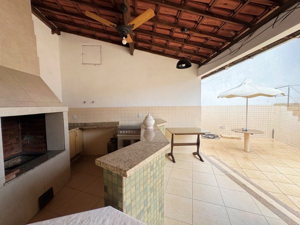 Área gourmet coberta com churrasqueira e bancada, com terraço e guarda-sol ao fundo
